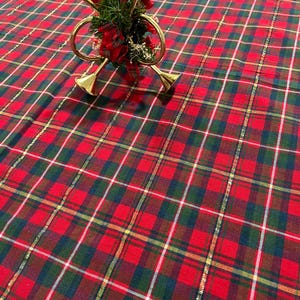 May include: A decorative ornament featuring three gold-toned miniature French horns arranged in a cluster, accented with green pine needles, small red berries, and a red plaid ribbon, all suspended by a thin red cord over a red, green, and gold plaid fabric.