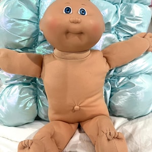 May include: A doll with a round, tan vinyl head, bright blue eyes, and a small tuft of light brown yarn hair. The doll has a soft, tan fabric body with a stitched navel and outstretched arms, featuring rosy cheeks and a neutral facial expression.