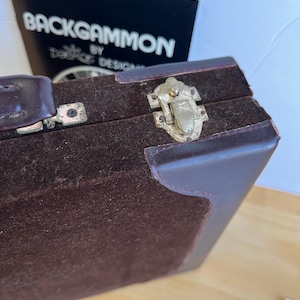 May include: A brown leather backgammon case with a metal latch. The case is lined with brown felt.