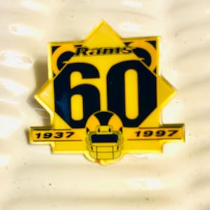 May include: A yellow and navy blue pin commemorating the 60th anniversary of the Rams football team. The pin features the number 60 in navy blue, the team name, and the years 1937 and 1997.