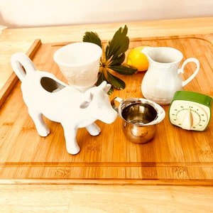 May include: A white ceramic cow creamer, a white ceramic pitcher, a stainless steel measuring cup, a green kitchen timer, and a lemon on a wooden cutting board.