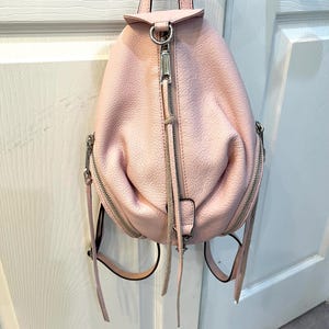 Op de afbeelding: Een lichtroze leren rugzak met zilverkleurige hardware. De tas heeft een afgeronde vorm met een handvat aan de bovenkant, een ritssluiting aan de voorkant en twee zijritsen. De rugzak heeft verstelbare bandjes.