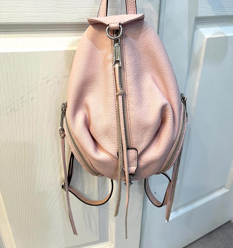 Op de afbeelding: Een lichtroze leren rugzak met zilverkleurige hardware. De tas heeft een afgeronde vorm met een handvat aan de bovenkant, een ritssluiting aan de voorkant en twee zijritsen. De rugzak heeft verstelbare bandjes.