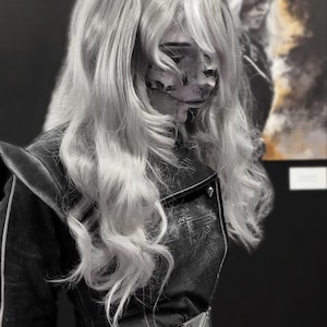 May include: A person with long, wavy, light-colored hair and a detailed face mask. They wear a dark jacket with a white belt and a silver buckle. A blurred image of the person is in the background.
