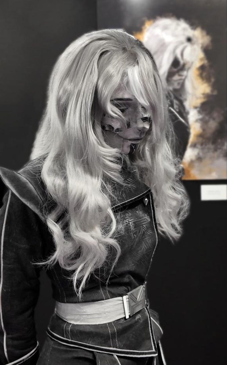 May include: A person with long, wavy, light-colored hair and a detailed face mask. They wear a dark jacket with a white belt and a silver buckle. A blurred image of the person is in the background.