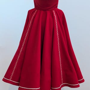 May include: A vibrant red cape with a high collar and a wide, flowing design. The cape features white stitching along the edges and seams, adding a decorative touch. The garment is displayed on a black stand, showcasing its full shape and details.