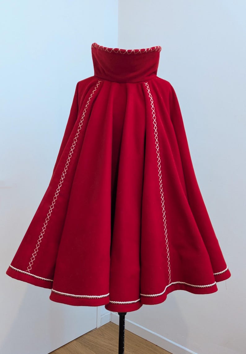 May include: A vibrant red cape with a high collar and a wide, flowing design. The cape features white stitching along the edges and seams, adding a decorative touch. The garment is displayed on a black stand, showcasing its full shape and details.
