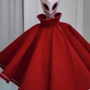May include: A red cape with white trim and a matching mask with horns. The mask is white with pink accents and eye holes. The cape has a high collar and white stitching.