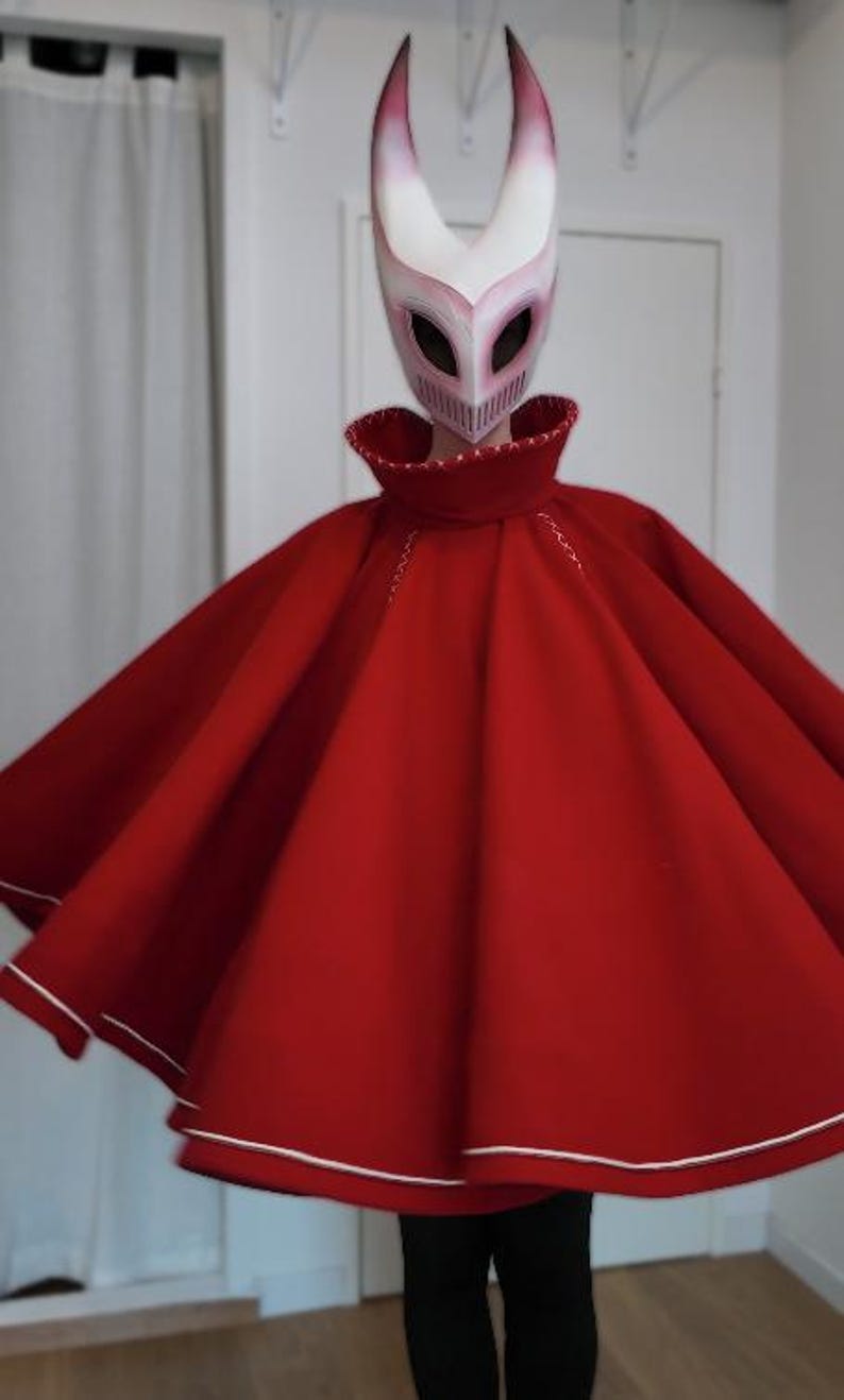 May include: A red cape with white trim and a matching mask with horns. The mask is white with pink accents and eye holes. The cape has a high collar and white stitching.