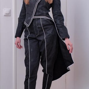 May include: A dark gray suit with white trim, consisting of a jacket and pants. The jacket has an asymmetrical design and the pants have a straight leg. The outfit is worn with white socks.