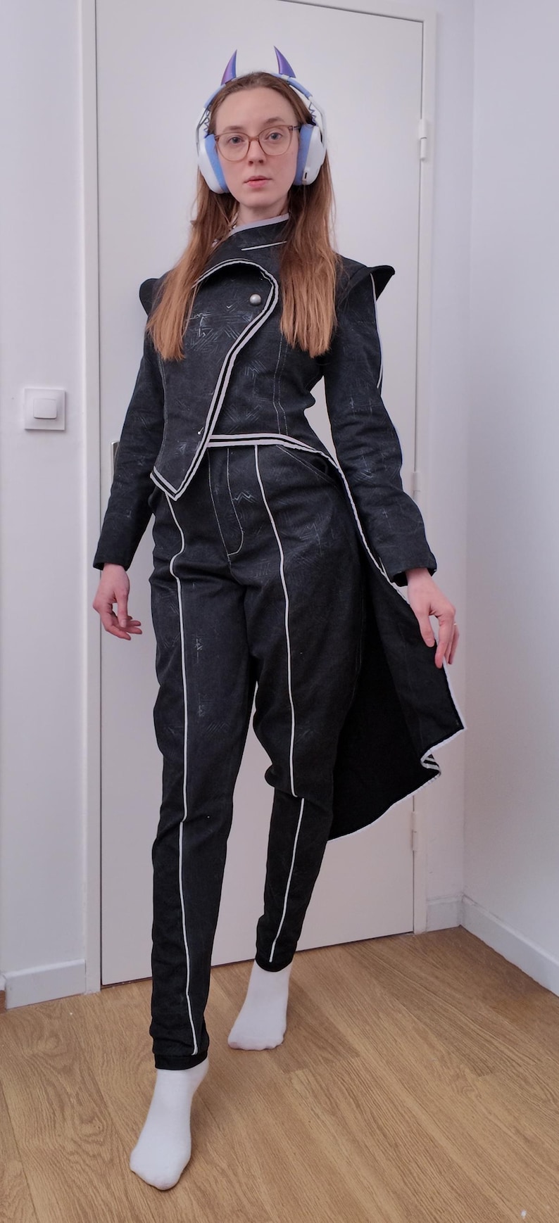 May include: A dark gray suit with white trim, consisting of a jacket and pants. The jacket has an asymmetrical design and the pants have a straight leg. The outfit is worn with white socks.