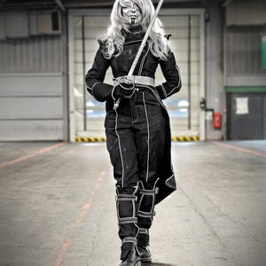 May include: A person in a black costume with white accents, holding a sword. The outfit includes a jacket, pants, and boots. The person has face paint and long blonde hair. The background is a large industrial space with a closed door.