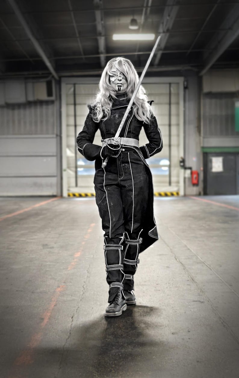 May include: A person in a black costume with white accents, holding a sword. The outfit includes a jacket, pants, and boots. The person has face paint and long blonde hair. The background is a large industrial space with a closed door.