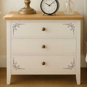 May include: A white wooden nightstand featuring three drawers with round bronze-toned knobs. The top and bottom drawers are decorated with black scrollwork corner accents. The nightstand has a light brown wood-grain top surface.