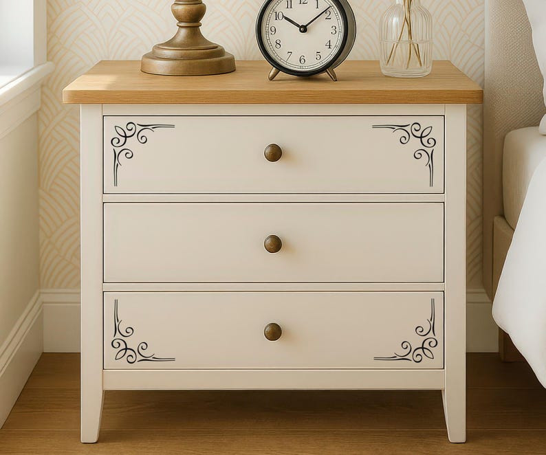 May include: A white wooden nightstand featuring three drawers with round bronze-toned knobs. The top and bottom drawers are decorated with black scrollwork corner accents. The nightstand has a light brown wood-grain top surface.