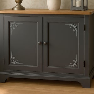 May include: A dark gray cabinet with two doors, featuring light gray decorative scrollwork in the corners of each door panel. The cabinet has a natural light wood-toned top surface and two small, round, dark gray knobs for opening the doors.