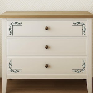 Può includere: Una cassettiera bianca a tre cassetti con un piano superiore in legno naturale e tre pomoncini rotondi marrone scuro. I cassetti superiore e inferiore sono decorati con motivi a volute grigio scuro sugli angoli laterali.