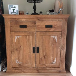 May include: A wooden cabinet with a medium brown finish, featuring two top drawers and two lower doors. Each door is accented with white decorative corner scrollwork. The unit includes rectangular black metal handles on all drawers and doors.