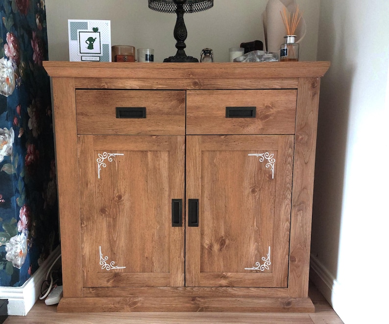 May include: A wooden cabinet with a medium brown finish, featuring two top drawers and two lower doors. Each door is accented with white decorative corner scrollwork. The unit includes rectangular black metal handles on all drawers and doors.