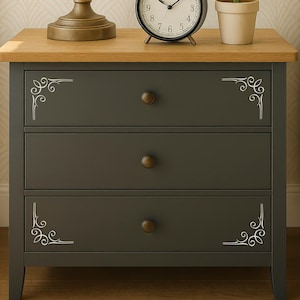 May include: A dark gray three-drawer nightstand with a light oak-colored wooden top. Each drawer features a small round bronze-colored knob and white decorative scrollwork patterns in the corners of the top and bottom drawer fronts.