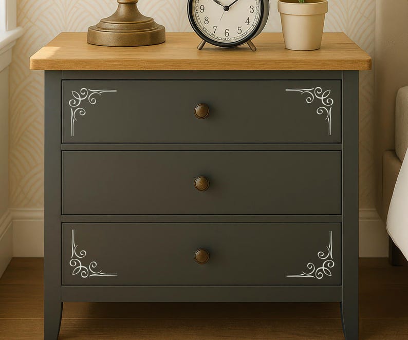 May include: A dark gray three-drawer nightstand with a light oak-colored wooden top. Each drawer features a small round bronze-colored knob and white decorative scrollwork patterns in the corners of the top and bottom drawer fronts.