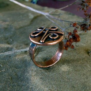 May include: A copper ring with a circular design featuring a stylized knot. The ring is on a textured gray surface.