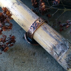 May include: A copper ring with the word "Strength" engraved on the band. The ring is resting on a piece of bamboo.