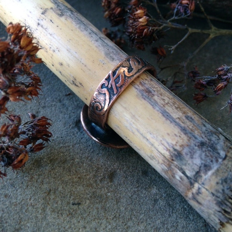 May include: A copper ring with the word "Strength" engraved on the band. The ring is resting on a piece of bamboo.