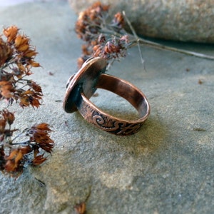May include: A copper ring with a textured band and a large oval-shaped stone set in the top. The ring is sitting on a gray, textured surface.