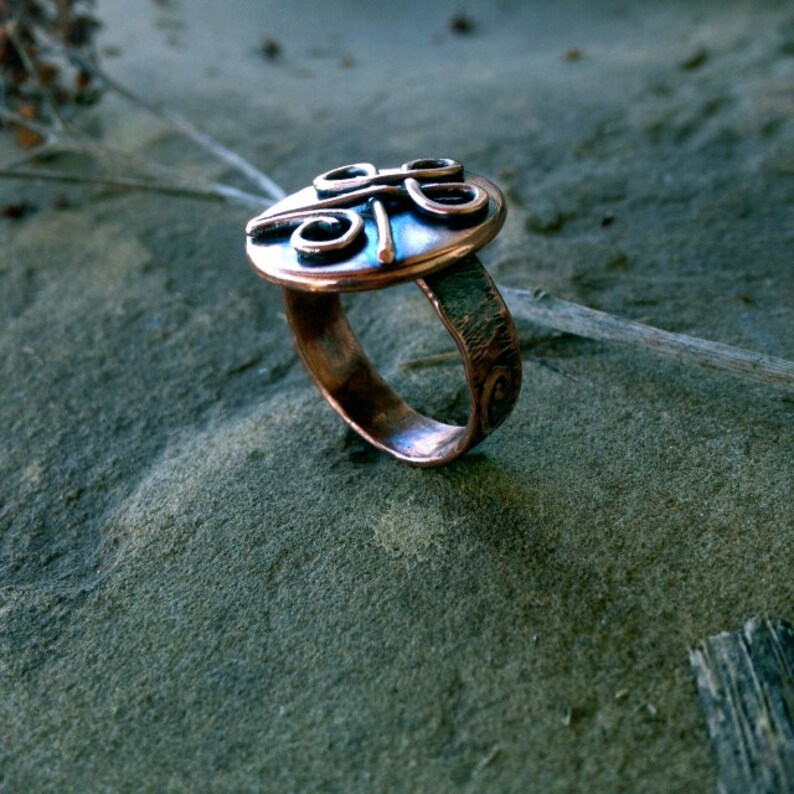May include: A copper ring with a textured band and a circular top featuring a stylized design of four overlapping circles.