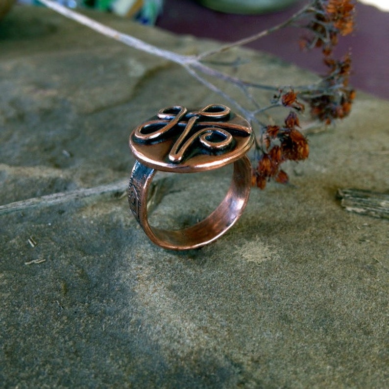 May include: A copper ring with a circular design featuring a stylized knot. The ring is on a textured gray surface.