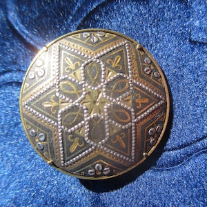 Peut inclure: Une broche ronde en argent et or avec un motif en étoile. La broche a un fond noir avec des accents argentés et dorés.