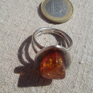 May include: A silver ring with a large, amber-colored stone. The ring has a double band design and a bezel setting for the stone. A Euro coin is next to the ring for size comparison.