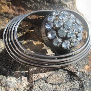 Puede incluir: Un broche de plata con un racimo de piedras de cristal azul claro dispuestas en forma circular. El broche está rodeado por una serie de círculos concéntricos.