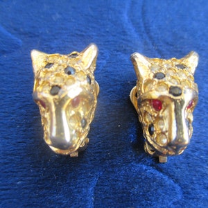 May include: A pair of gold-tone clip-on earrings featuring a leopard head design with black and clear rhinestones and red accents.