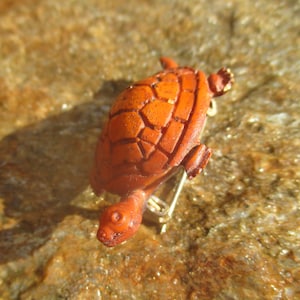 May include: A small, orange turtle figurine with a detailed shell. The turtle is made of wood and has a gold-colored pin on the back.