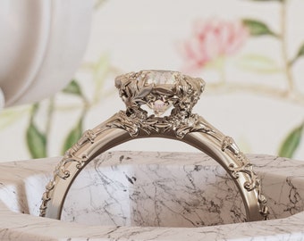 Könnte beinhalten: Ein silberner Ring mit einem großen, klaren Edelstein, der in einem zarten, floralen Design gefasst ist. Der Ring wird auf einer weißen Marmorfläche präsentiert. Der Text "Stellar Fields Heirlooms" ist im Hintergrund sichtbar.