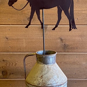 May include: A metal silhouette of a cowgirl riding a horse, mounted on a metal rod, inserted into a vintage metal milk jug.