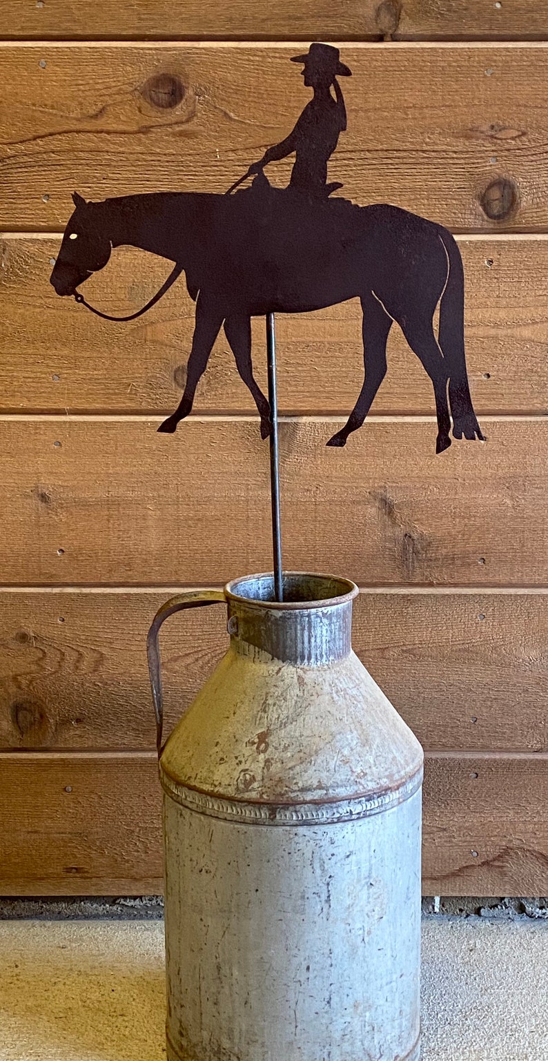 May include: A metal silhouette of a cowgirl riding a horse, mounted on a metal rod, inserted into a vintage metal milk jug.