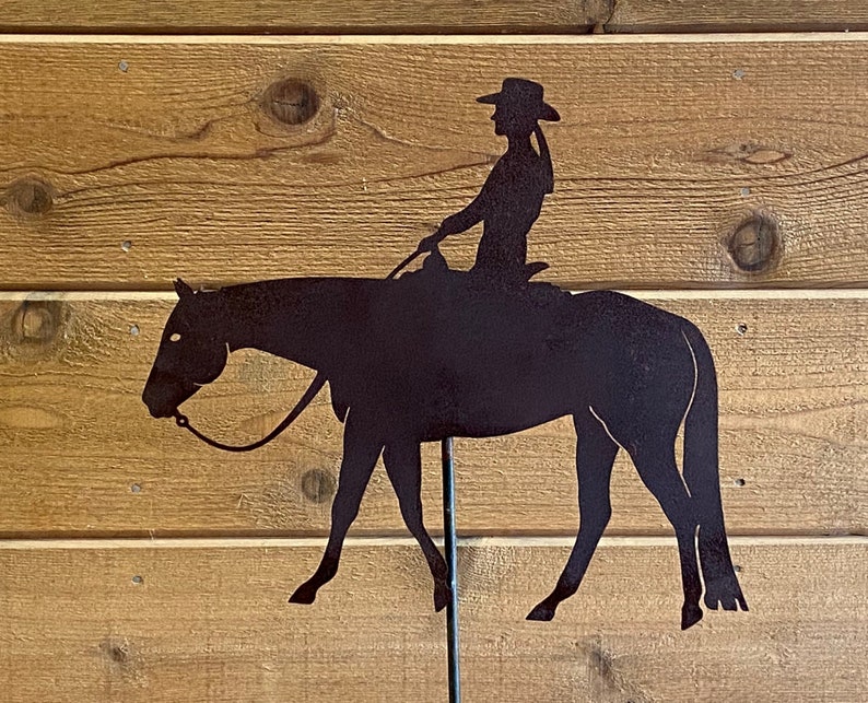 May include: A black metal silhouette of a person riding a horse. The silhouette is mounted on a metal stake.