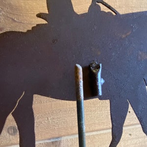 May include: A metal silhouette of a horse and rider, with a rusty brown finish. The silhouette is mounted on a metal rod, which is inserted into a wooden surface.