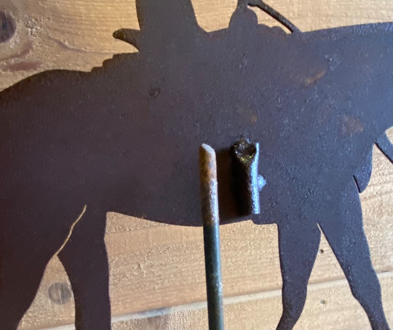 May include: A metal silhouette of a horse and rider, with a rusty brown finish. The silhouette is mounted on a metal rod, which is inserted into a wooden surface.
