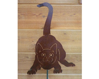 May include: A rusty brown metal silhouette of a cat with its tail curled up. The cat is facing forward with its head tilted down and its eyes looking up. The silhouette is mounted on a green metal stake.
