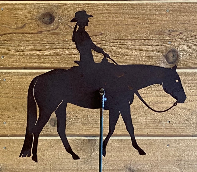 May include: Silhouette of a person riding a horse, cut from metal and mounted on a metal stand. The silhouette is black and the stand is silver.