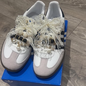 Op de afbeelding: Witte sneakers met zwarte strepen en een lichtgrijze neus. De sneakers zijn versierd met kant, parels en zwarte en gouden kralen. De tekst "Crystally Boutique" is zichtbaar op de schoen.