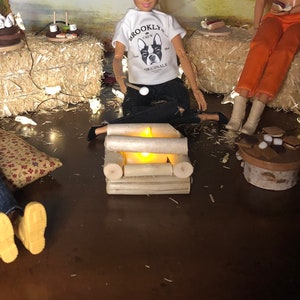 May include: Three dolls are gathered around a miniature campfire made of logs. The dolls are wearing casual clothing and are sitting on hay bales. The dolls are holding sticks and marshmallows. The scene is lit by string lights.