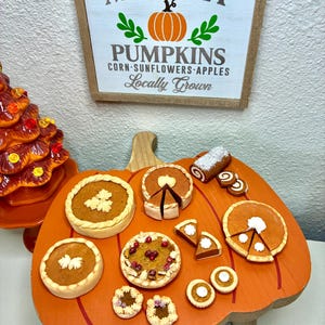Puede incluir: Una colección en miniatura de postres de arcilla con temática otoñal, incluyendo pasteles de calabaza, tartas y un brazo de gitano sobre una bandeja de madera con forma de calabaza, bajo un letrero que dice Harvest MARKET PUMPKINS.