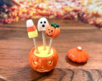 Könnte beinhalten: Ein kleiner, orangefarbener Kürbisbehälter mit abnehmbarem Deckel, der drei Holzspieße mit kleinen Figuren enthält: ein weiß-gelb-oranges Zuckermais-Bonbon, ein weißer Geist mit schwarzen Augen und ein kleiner orangefarbener Kürbis.