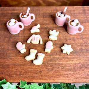 May include: Miniature pink mugs of hot chocolate with marshmallows and straws, alongside tiny cookies shaped like mittens, boots, a sweater, a hat, an angel, and a snowflake. The items are arranged on a wooden surface, evoking a cozy, festive theme.