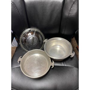 May include: A set of two silver-toned metal cooking pans with side handles and a single clear glass lid featuring a central handle. The pans have a smooth, metallic interior finish and a circular shape, designed for stovetop or oven use.